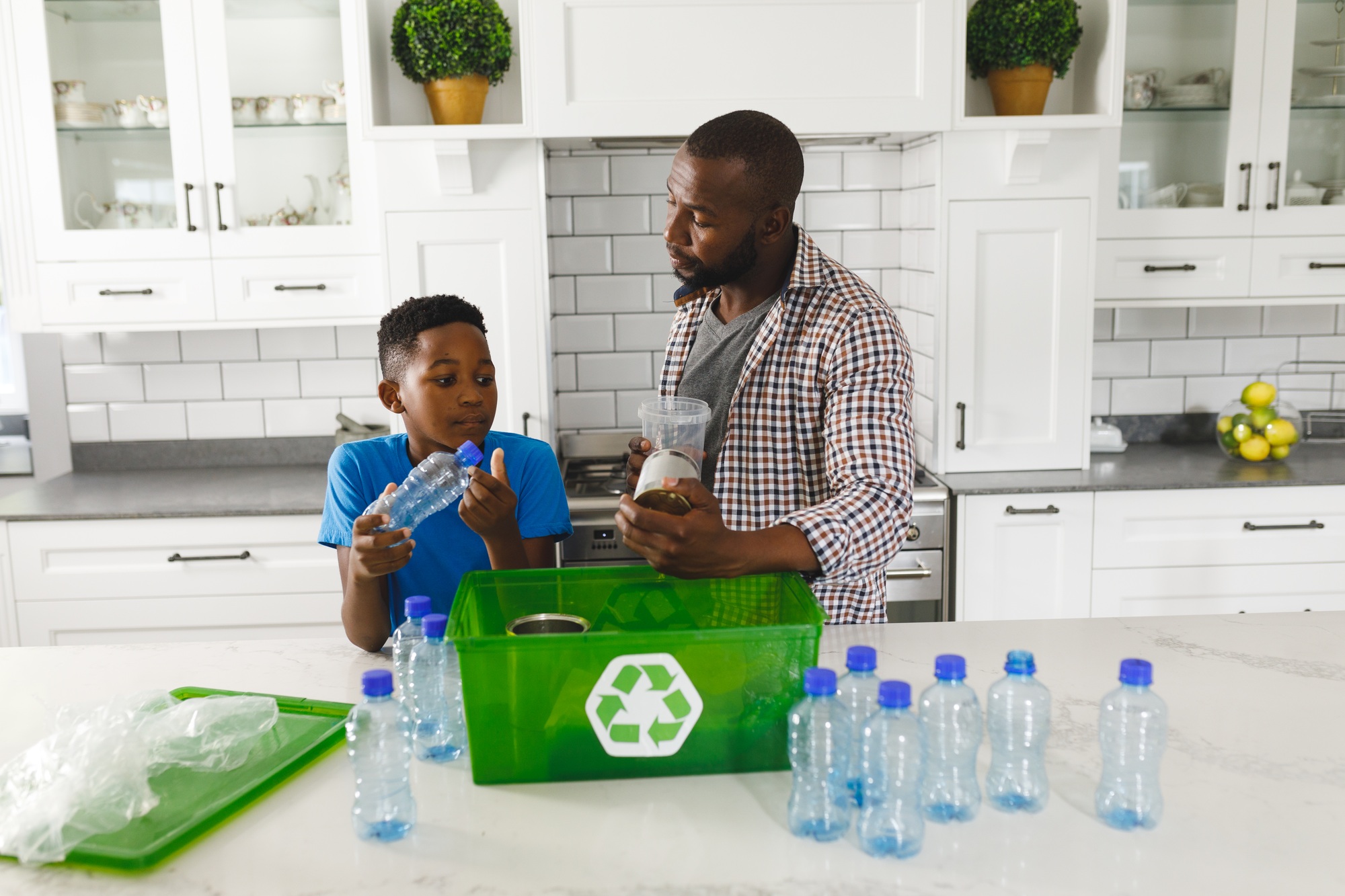 Happy african american father and son in kitchen talking and sorting plastic waste for recycling